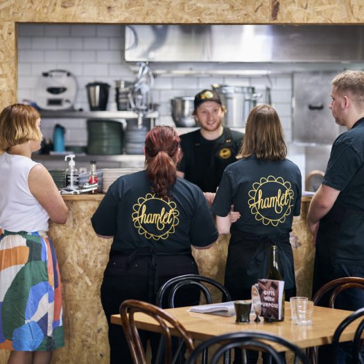 Several people standing at a cafe kitchen window, listening to a chef talking